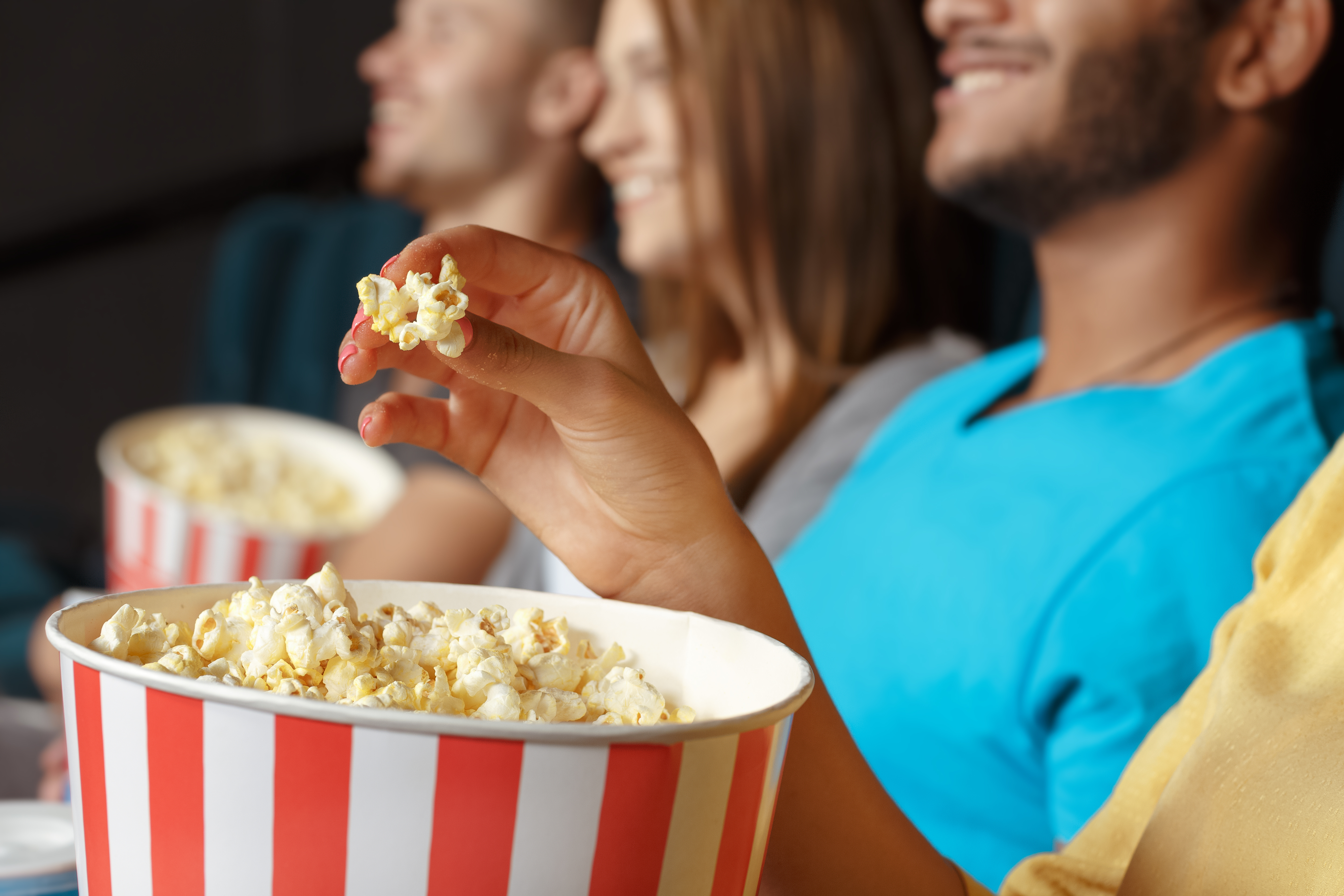 People are in a theater, smiling and eating popcorn. The fingers of a person are holding a piece of popcorn above a large popcorn container