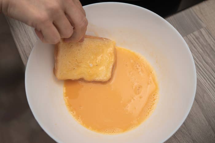 A hand dips a slice of bread into a bowl containing a beaten egg mixture, preparing to make French toast