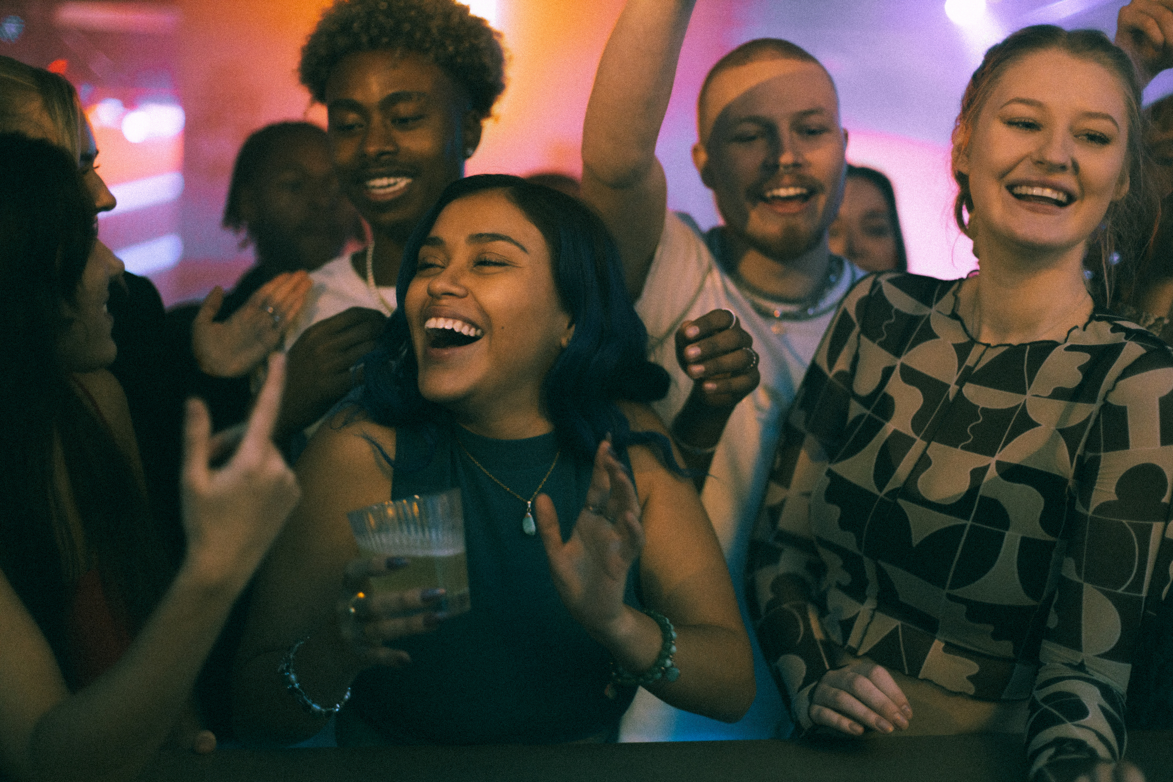 A joyful group of young adults, including a smiling woman in a patterned top, celebrate together in a lively, intimate party setting