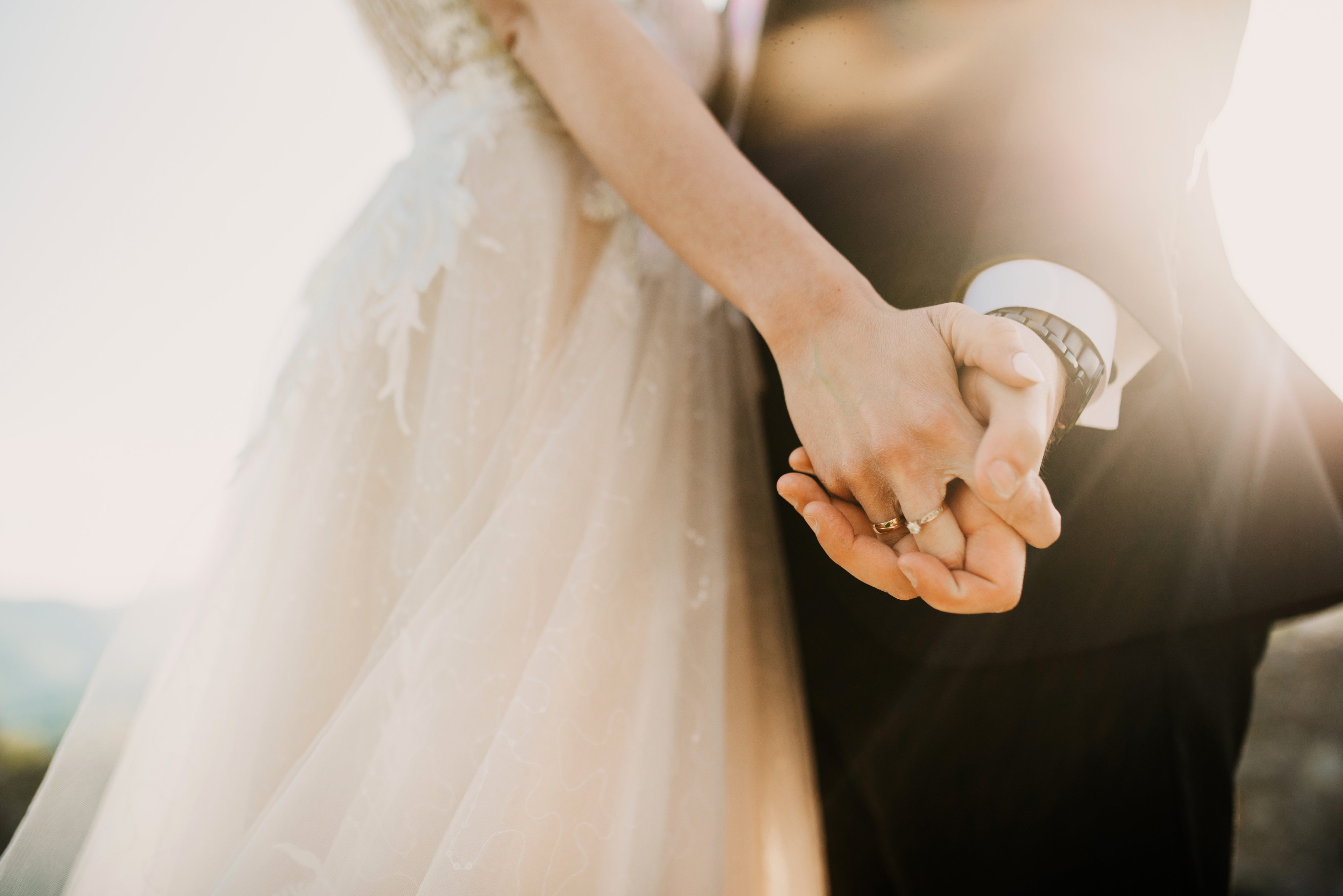A bride and groom hold hands tightly, showcasing their wedding rings. The bride&#x27;s lacy dress and the groom&#x27;s suit sleeve with cufflinks are visible