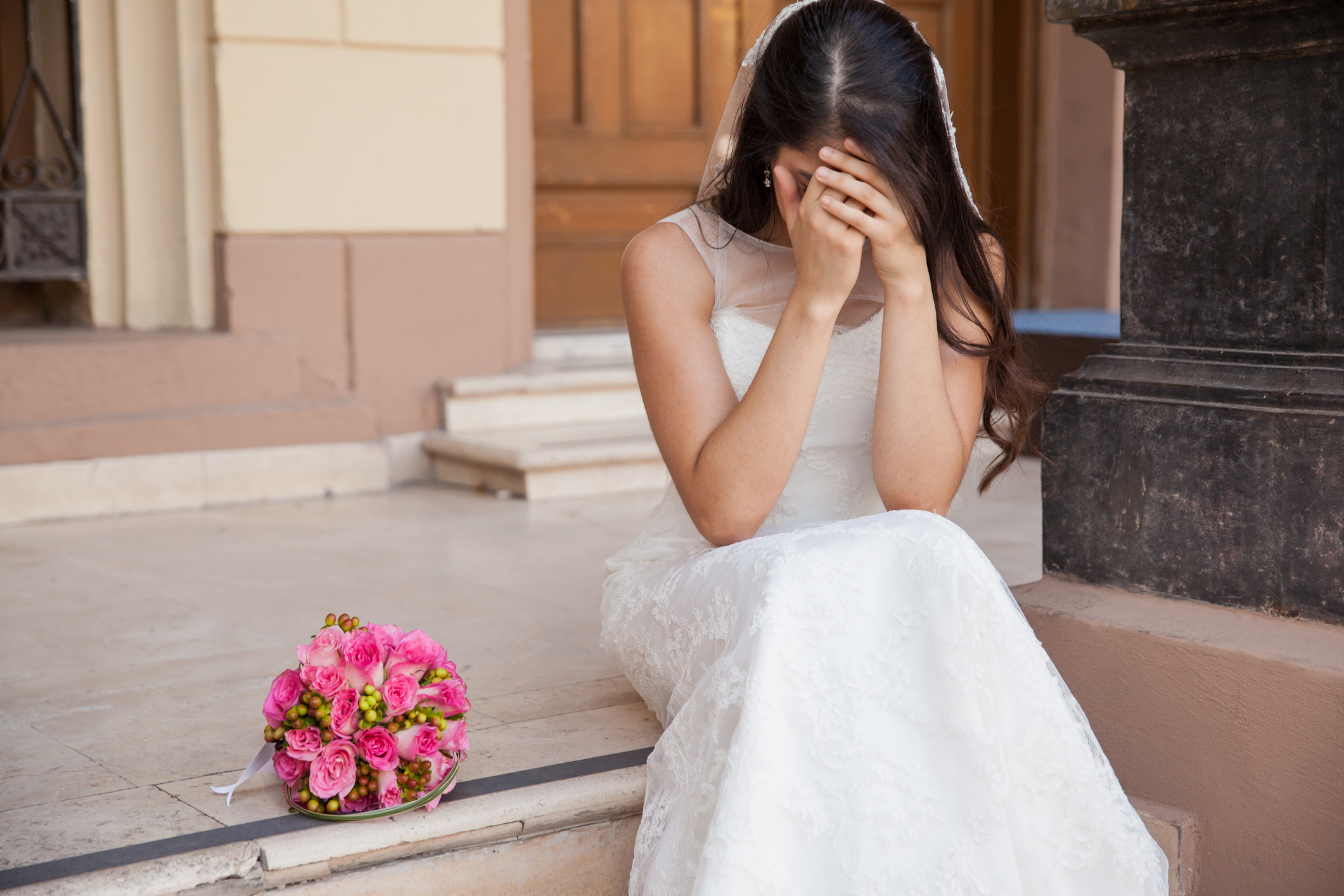 A bride in a white lace wedding dress sits on a step, covering her face with her hands. A bouquet of flowers is placed beside her