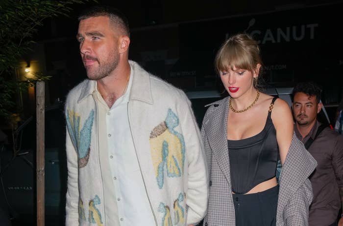 Travis Kelce (left) in a patterned jacket and Taylor Swift (right) in a corset top and checkered coat, walking together outside at night