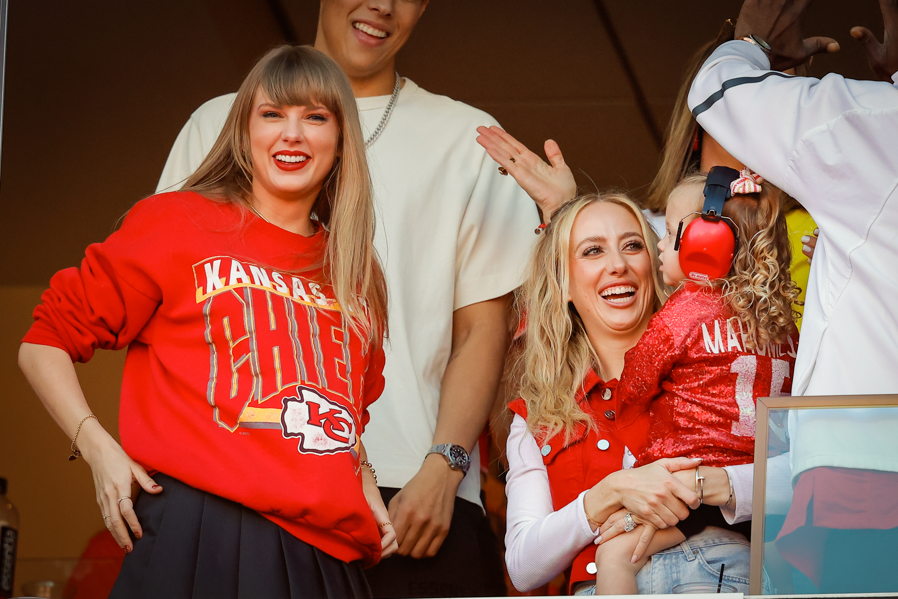Taylor Swift and Brittany Mahomes smiling and enjoying a sports event. Brittany is holding a young child with headphones, surrounded by others cheering