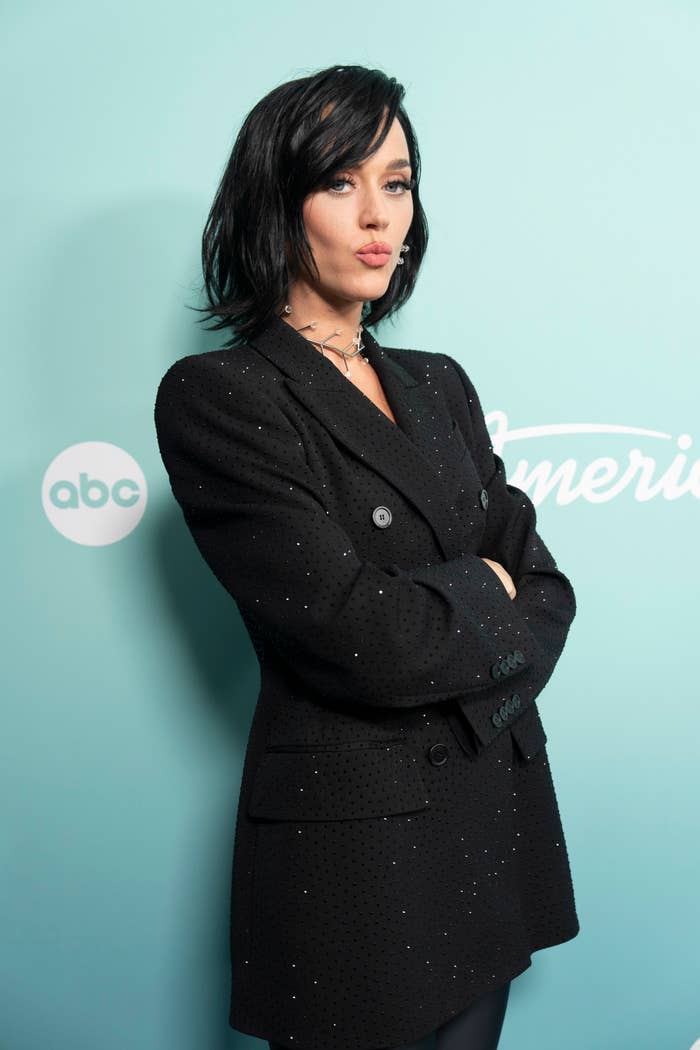 Katy Perry poses with crossed arms in a chic, dark, oversized blazer adorned with subtle sparkles at an "American Idol" event