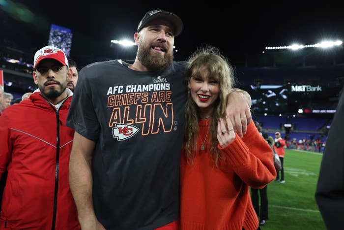 Travis Kelce and Taylor Swift are on a football field. Travis is wearing a "AFC Champions Chiefs Are All In" T-shirt, and Taylor is in a sweater