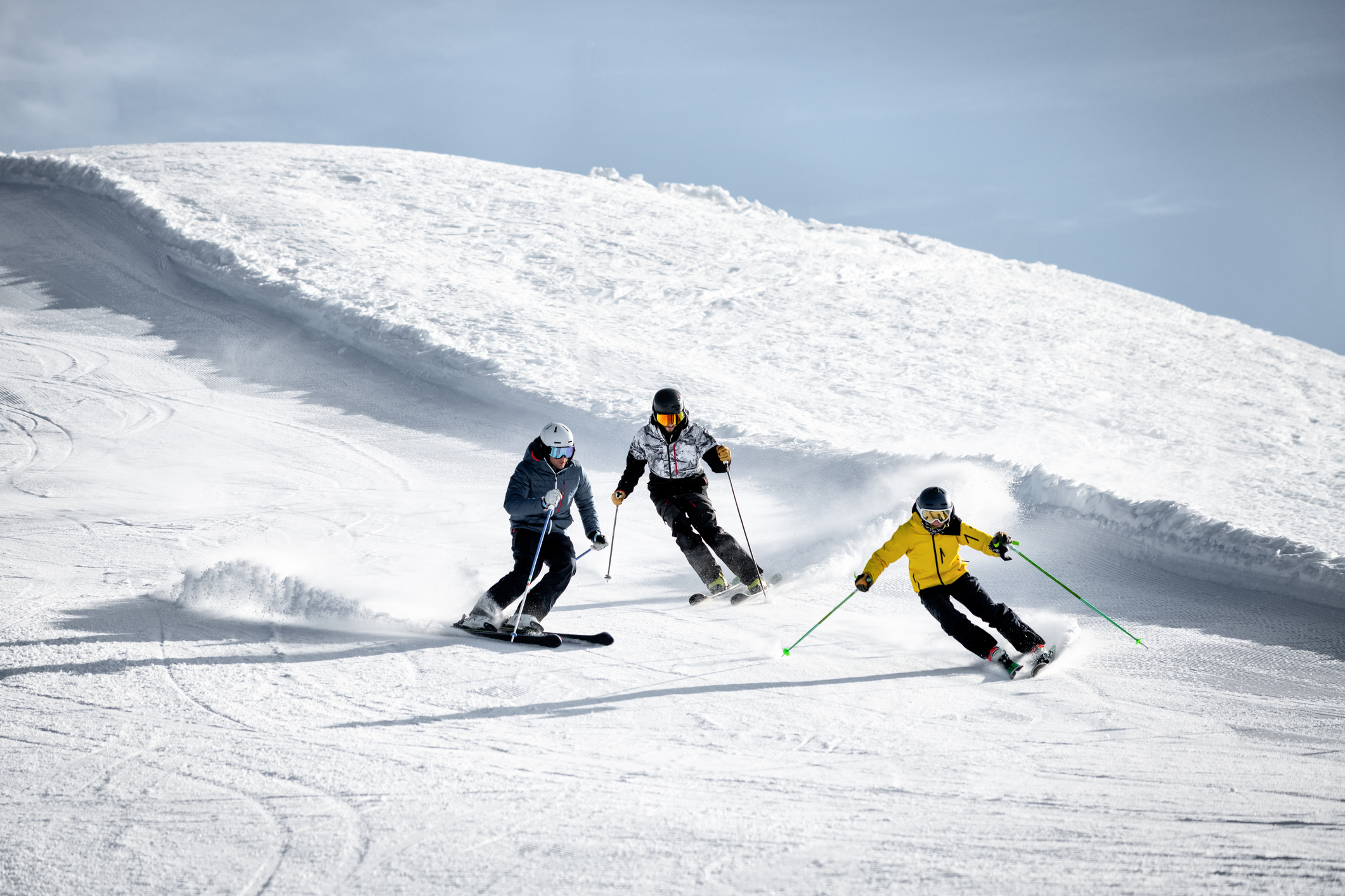 Three skiers navigate a snowy slope, creating spray as they turn. They are dressed in winter gear, with one skier in a yellow jacket leading the group