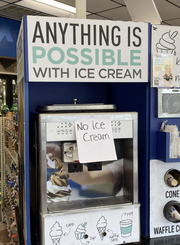 Soft serve ice cream machine at a shop with a "No Ice Cream" sign taped to it. Surrounding signs read "Anything is possible with ice cream" and show different cone options