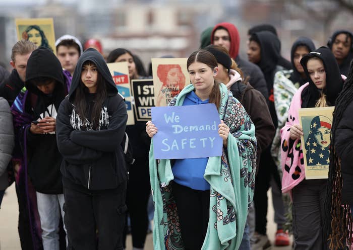 A group of people, mainly young women including a girl holding a sign that reads &quot;We Demand Safety,&quot; are participating in a protest