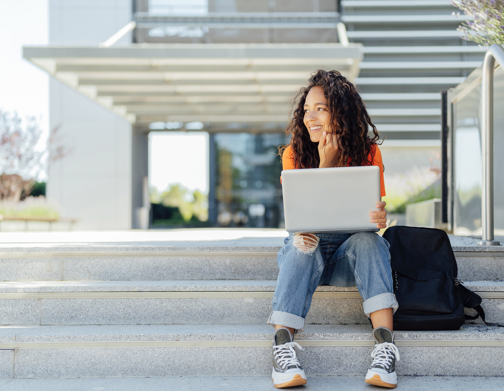 A woman sits on steps, smiling while using a laptop, with a backpack beside her. She wears a casual outfit of ripped jeans, sneakers, and a T-shirt