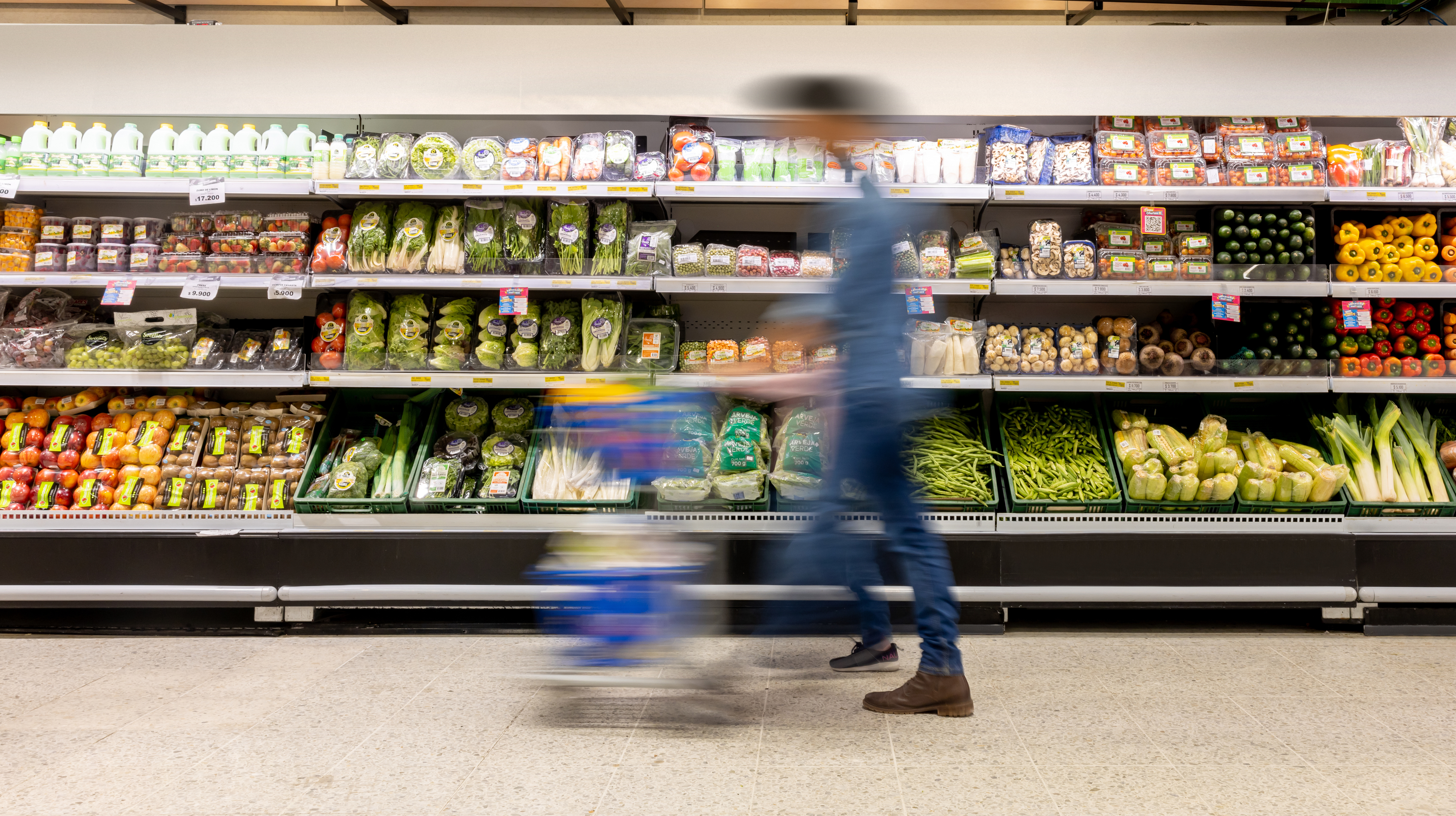 A person with a blurred motion pushes a shopping cart past a supermarket produce aisle stocked with various fruits, vegetables, and packaged goods