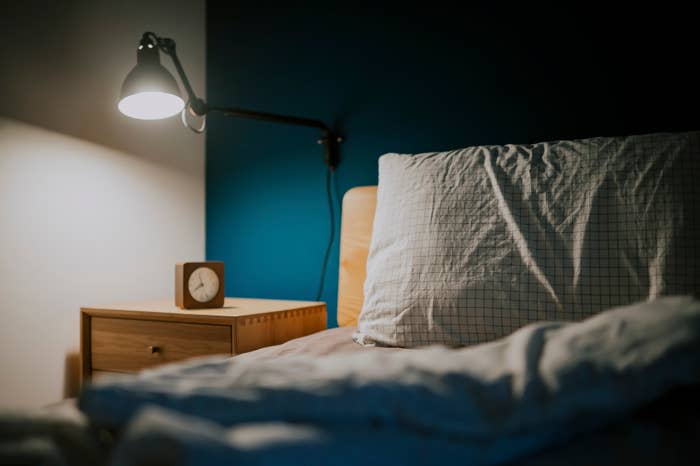 A lamp, nightstand, a modern clock, and corner of a bed with a white pillow on top