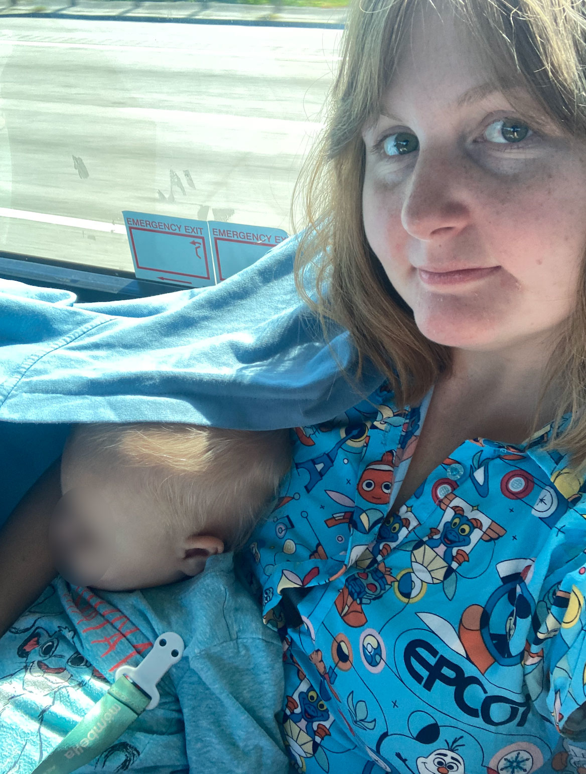 Woman in a blue shirt featuring Epcot and Disney characters sits on a bus with a child sleeping on her lap