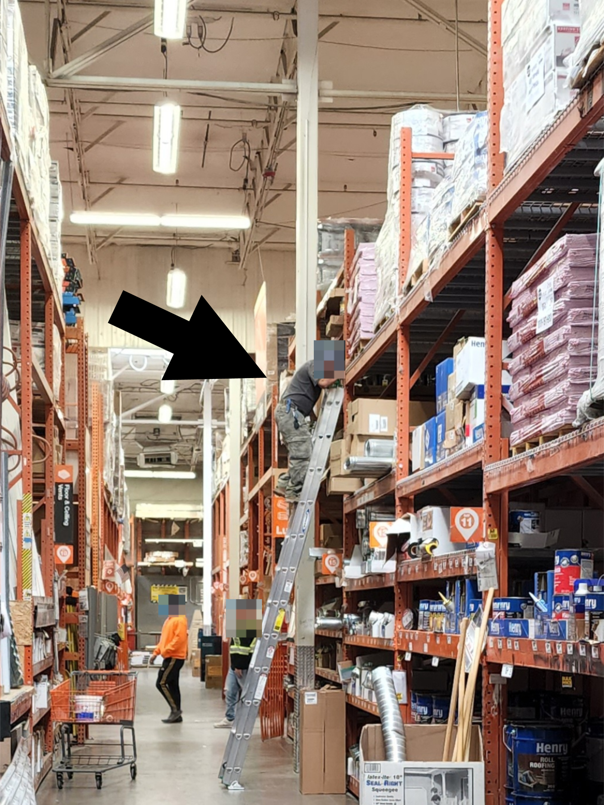 A worker on a ladder reaches for an item on a high shelf in a home improvement store, while another employee in an orange shirt pushes a cart nearby