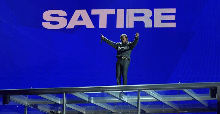 Rapper Kendrick Lamar performs on top of a glass box containing a car during a concert. &quot;SATIRE&quot; is written in large letters above him