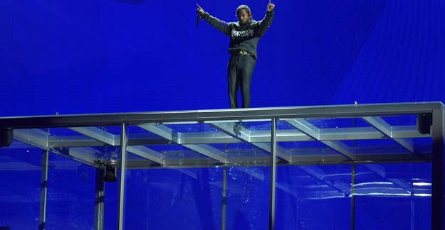 Rapper Kendrick Lamar performs on top of a glass box containing a car during a concert. "SATIRE" is written in large letters above him