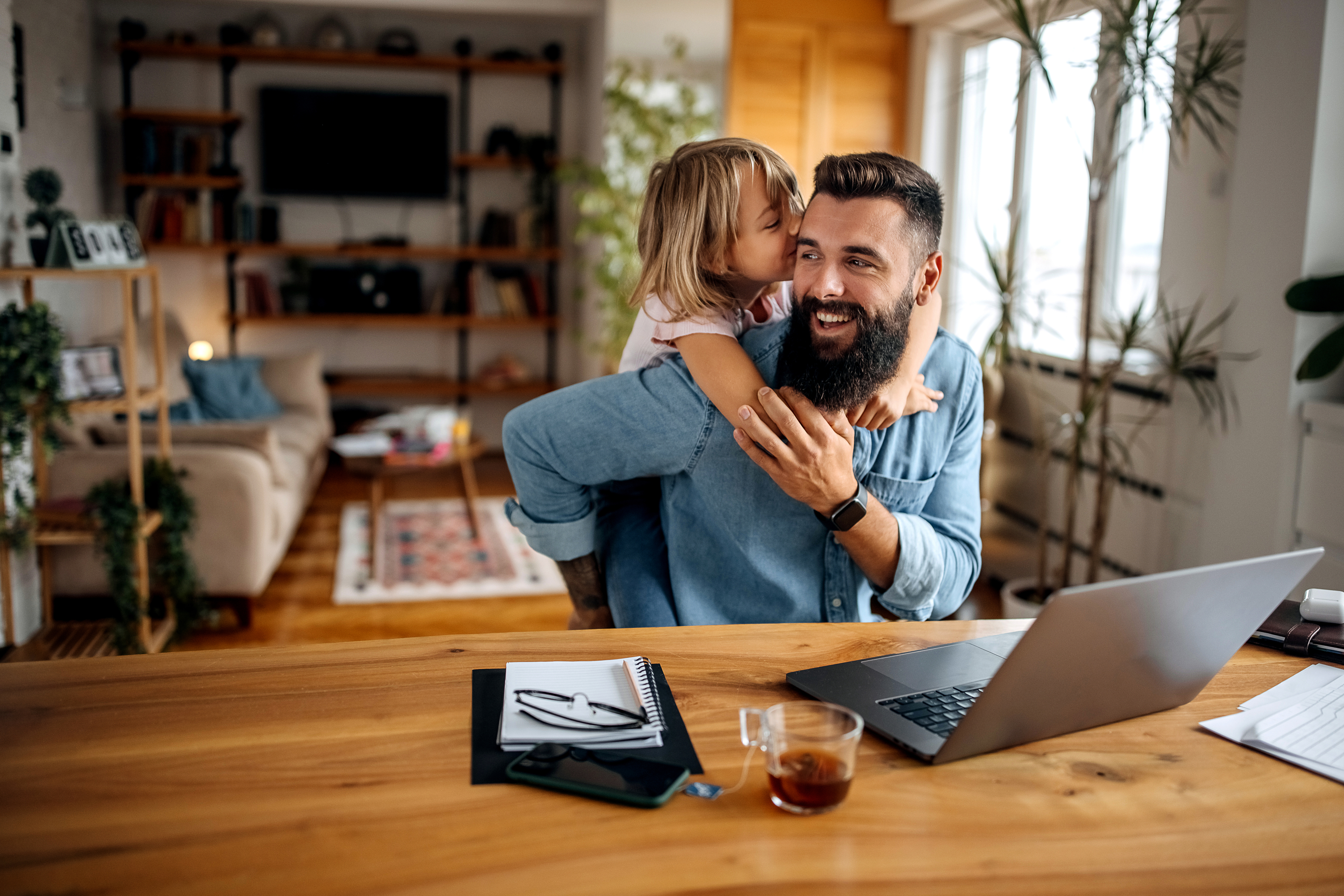 A bearded man is working from home on a laptop while a young child playfully kisses him on the cheek. Papers and a drink are on the wooden table