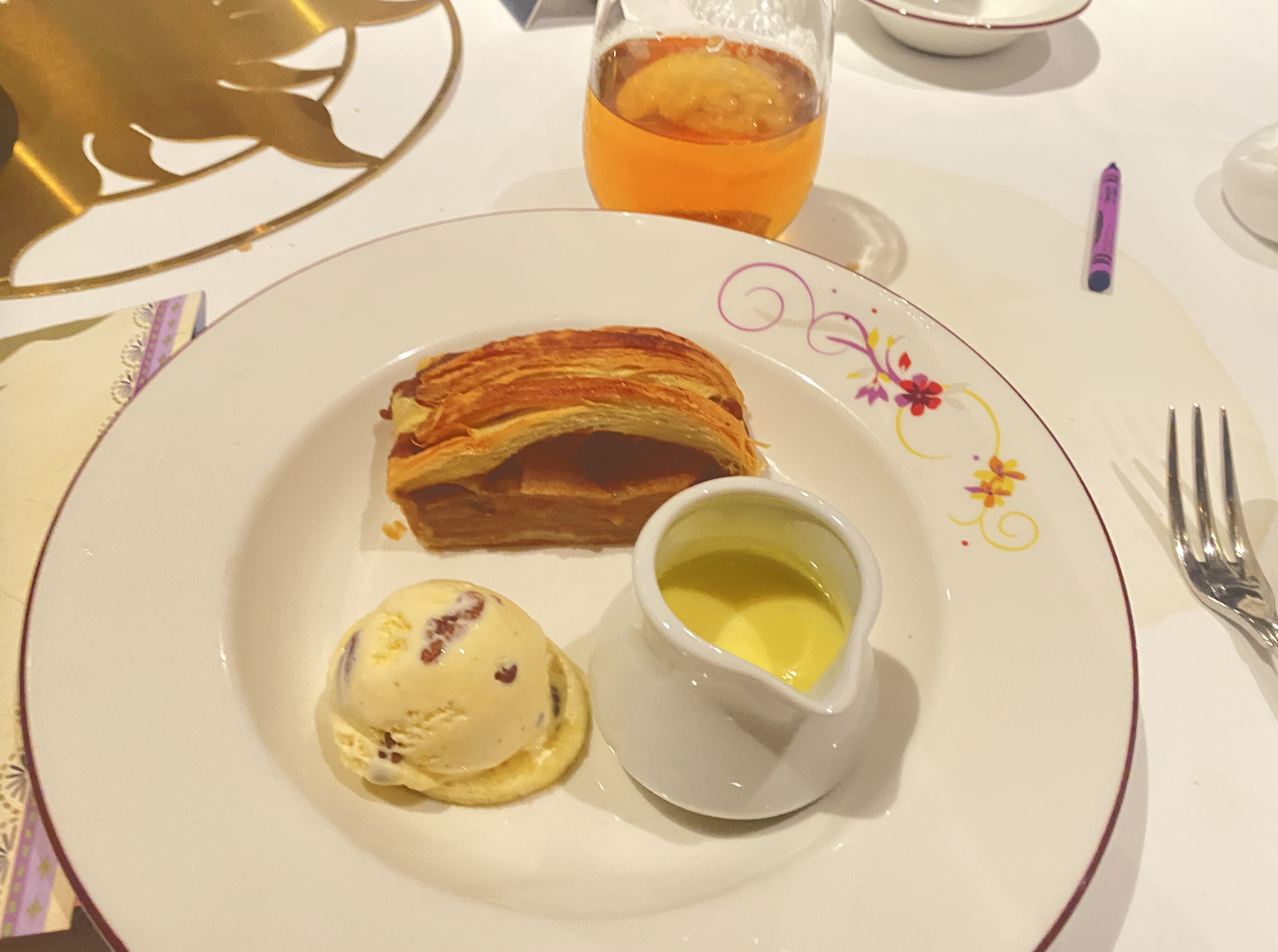 Dessert plate with a slice of layered pastry, a scoop of vanilla ice cream, and a small jug of yellow sauce, on a decorated table setting with a drink