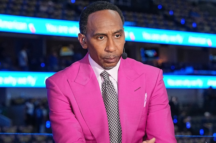 Stephen A. Smith seated, wearing a pink suit and polka dot tie, with a microphone headset in a sports arena background