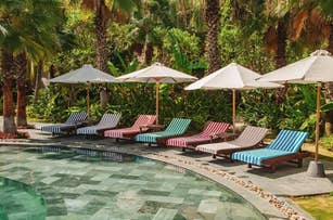 Poolside lounge chairs with striped cushions beneath umbrellas, surrounded by lush greenery in a tropical setting. Ideal for a shopping feature on outdoor furniture