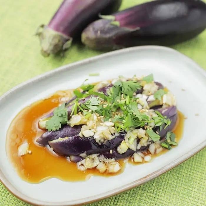 お皿に盛られたナスの料理、上には刻んだ香草とソースがかかっている。背景にはそのままのナスが見える。