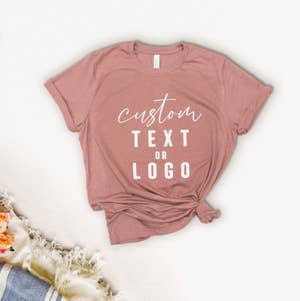 A pink t-shirt with the text "custom text or logo" displayed. The shirt is tied at the bottom right. A fuzzy blanket with flowers is partially visible in the corner