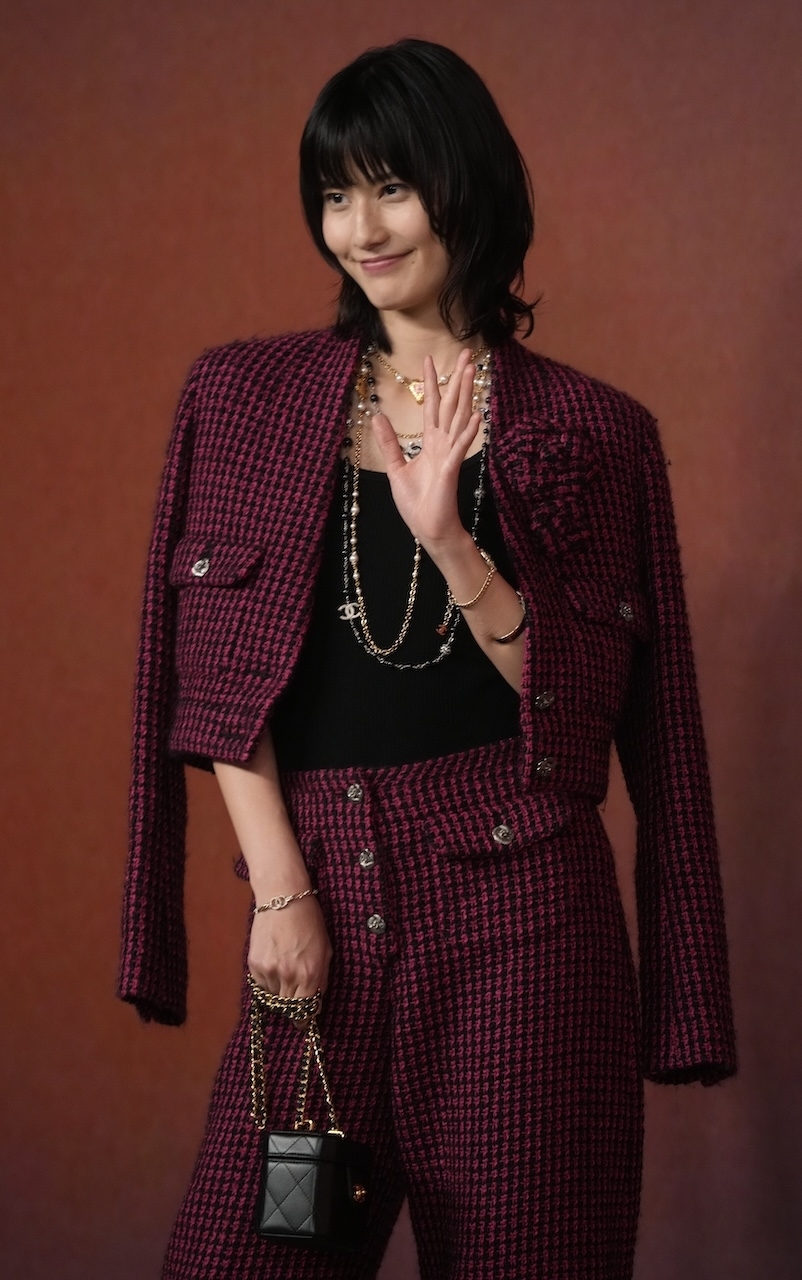 女性がカメラに向かって笑顔で手を振っている。彼女はカジュアルなツイードジャケットとパンツのセットを着て、ネックレスを身に着けている。名前は分かりません。