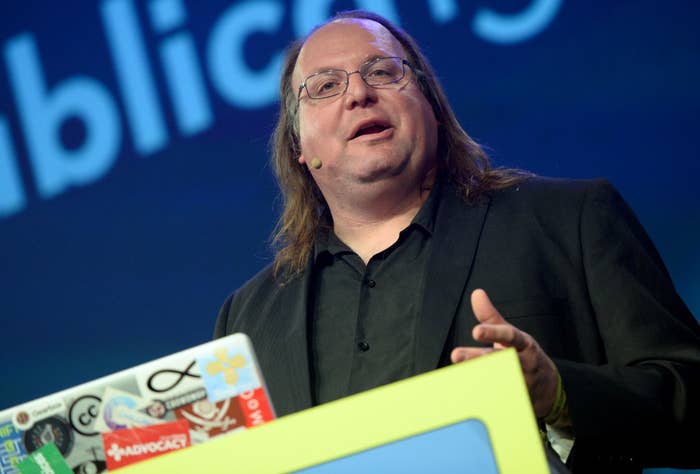 A man in a black suit, speaking on stage behind a laptop covered in various stickers