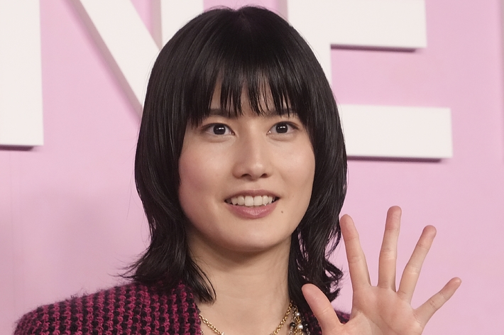 女性が笑顔で手を振っている。カメラを見つめ、ピンクの背景の前に立っている。