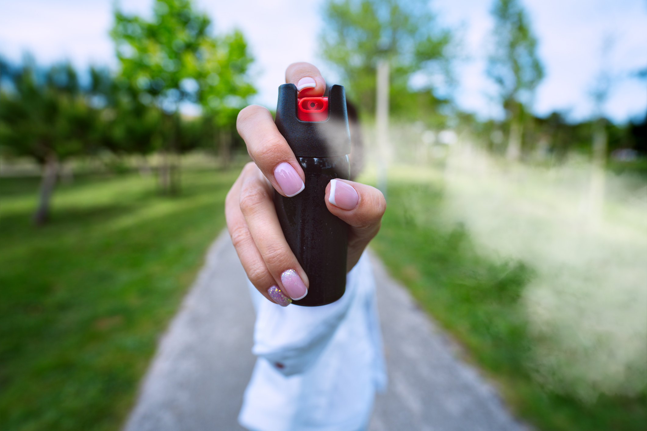 A person holding and spraying a can of pepper spray towards the camera in an outdoor park setting