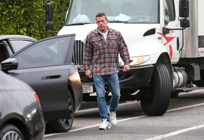 Ben Affleck in a casual plaid shirt, jeans, and sneakers, walking on a street near cars and a truck