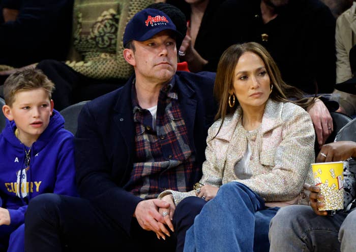 Jennifer Lopez and Ben Affleck sit courtside at a basketball game, with Affleck in a casual jacket and cap and Lopez in a chic coat. A child sits next to them