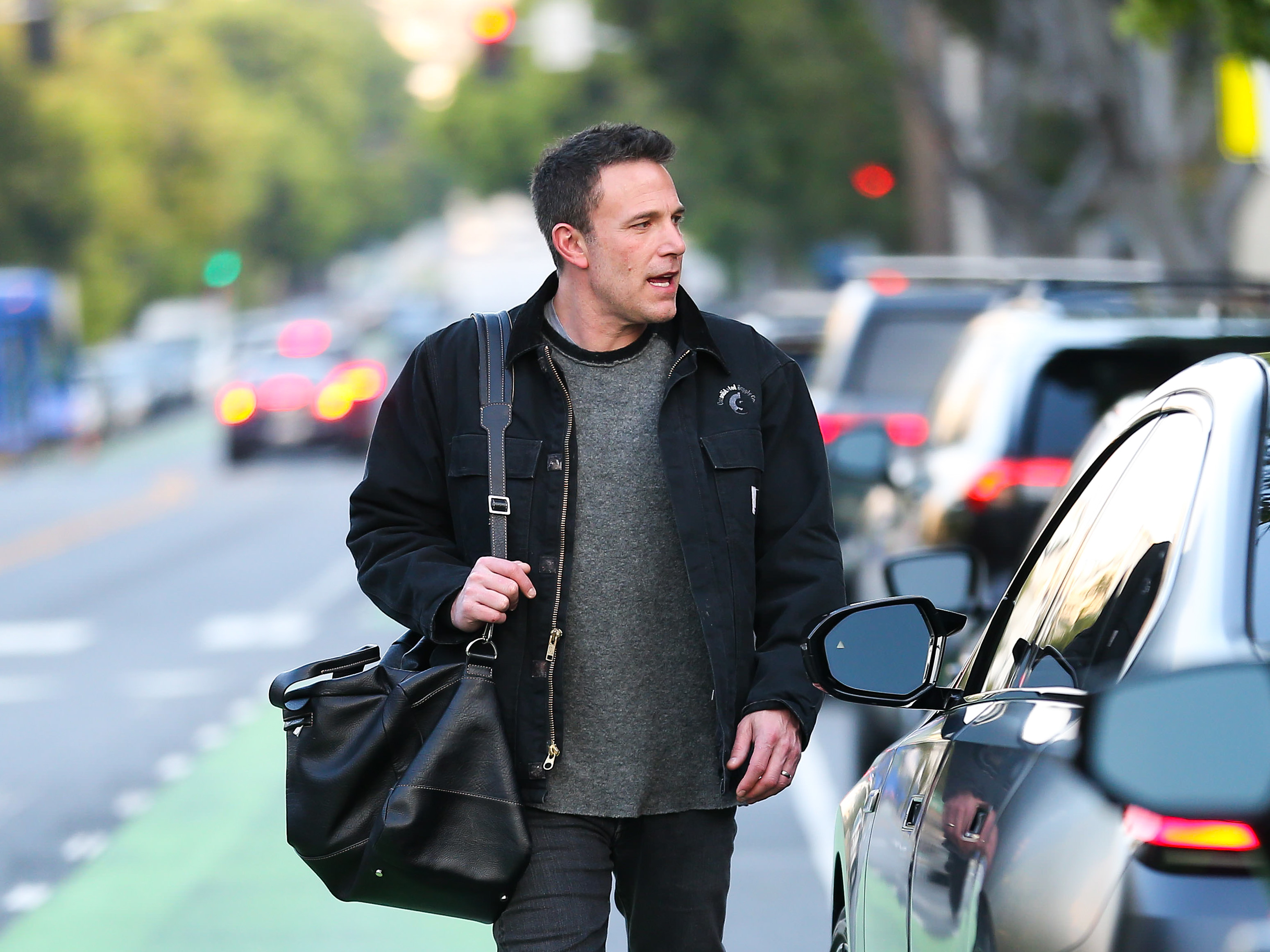 Ben Affleck walks on a city street wearing casual attire, carrying a large black bag, and looking towards a car