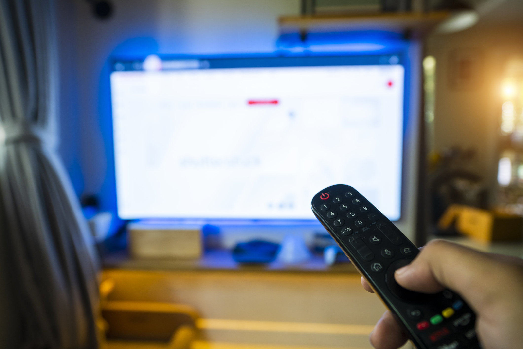 A hand holding a TV remote control is pointed towards a blurred TV screen in a living room setting
