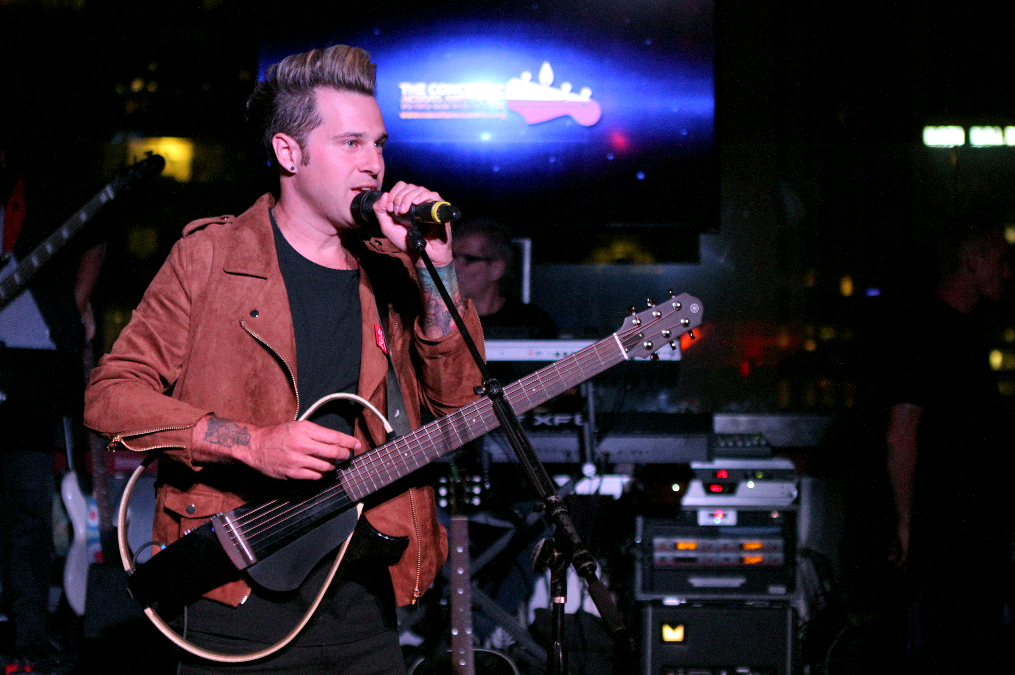 Ryan Cabrera performs on stage, holding a guitar and singing into a microphone. Musical equipment is visible in the background.