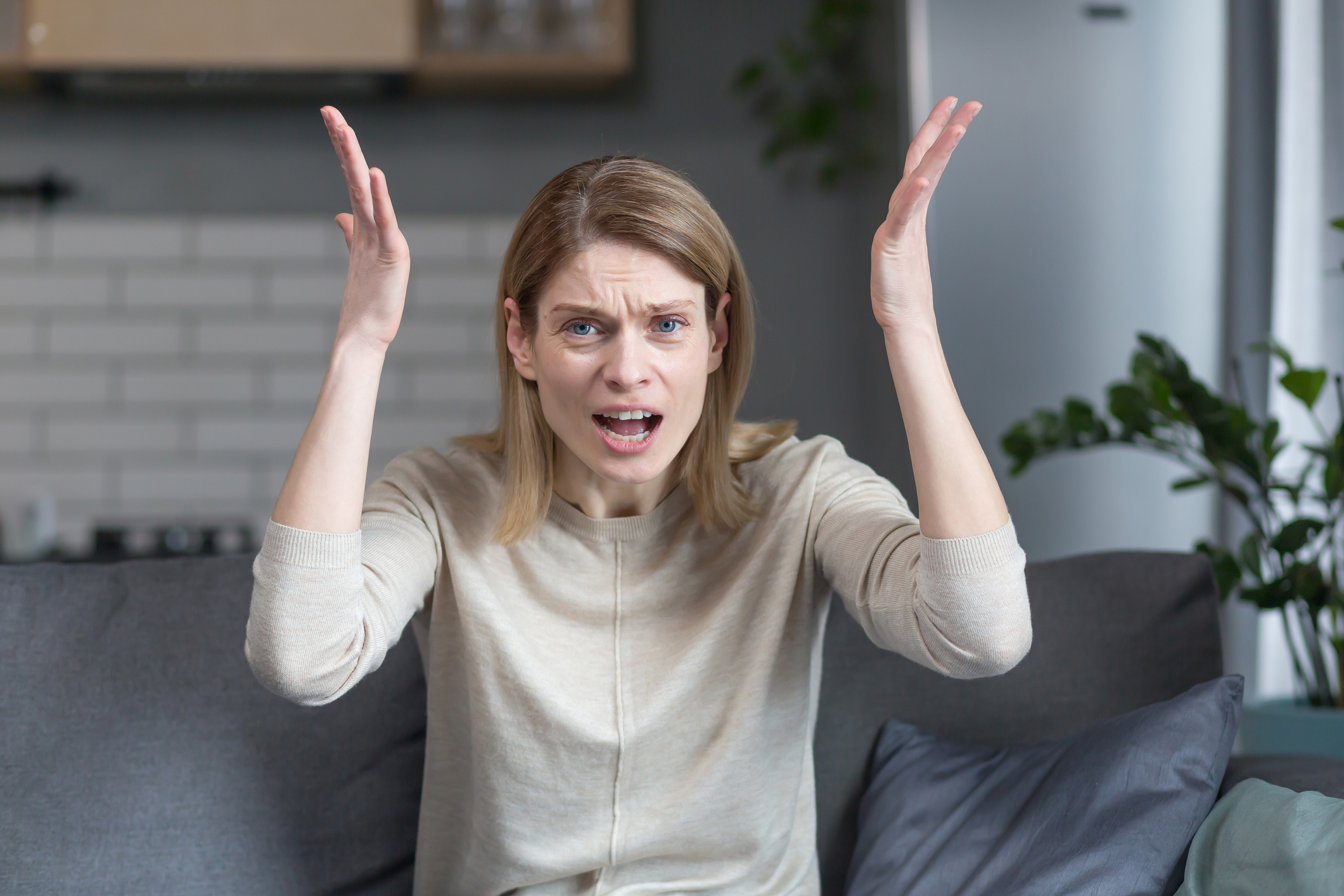 A woman with blonde hair and a beige top sits on a couch, looking frustrated with her hands raised