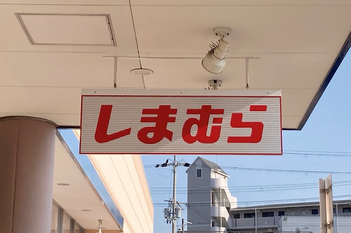 A sign with Japanese characters reads &ldquo;しまむら&rdquo; hanging from a building ceiling, with another building visible in the background