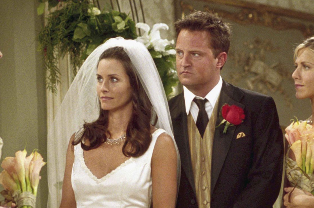 Courteney Cox in a wedding dress stands next to Matthew Perry in a suit with a red rose, and Jennifer Aniston by his side, in a wedding setting with flowers
