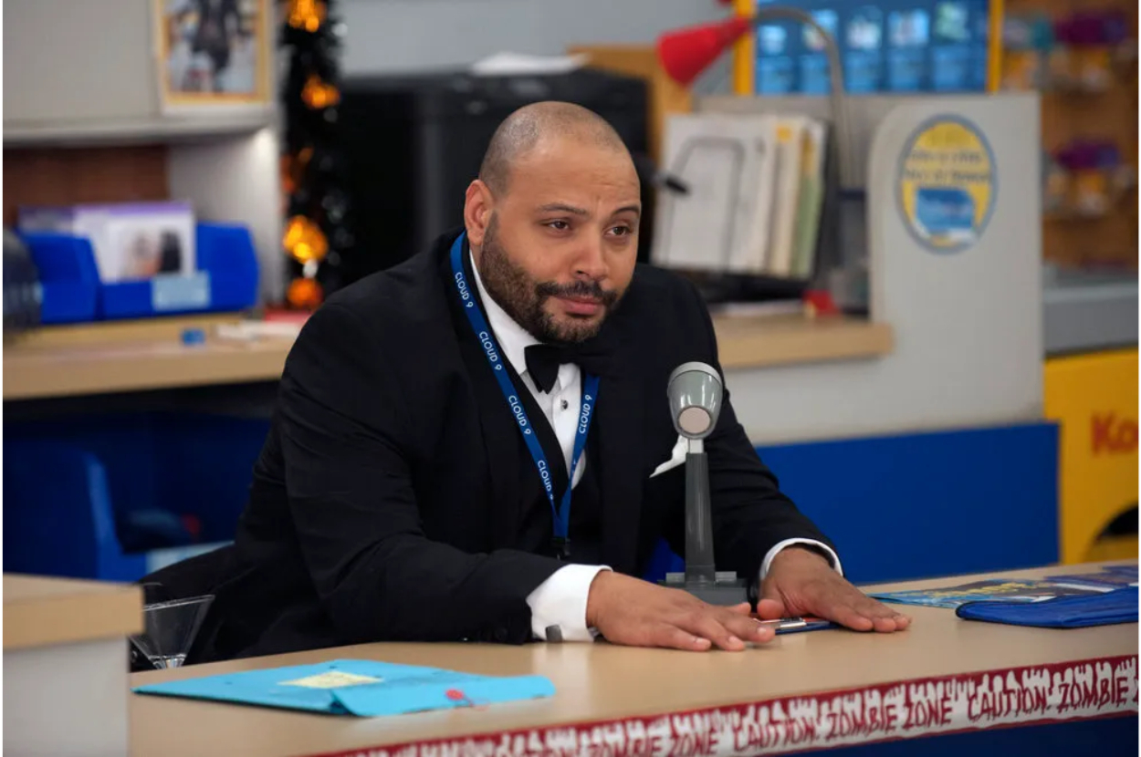 A man in formal wear with a coat and tie sits behind a desk with a microphone, appearing to address an audience or engage in a discussion