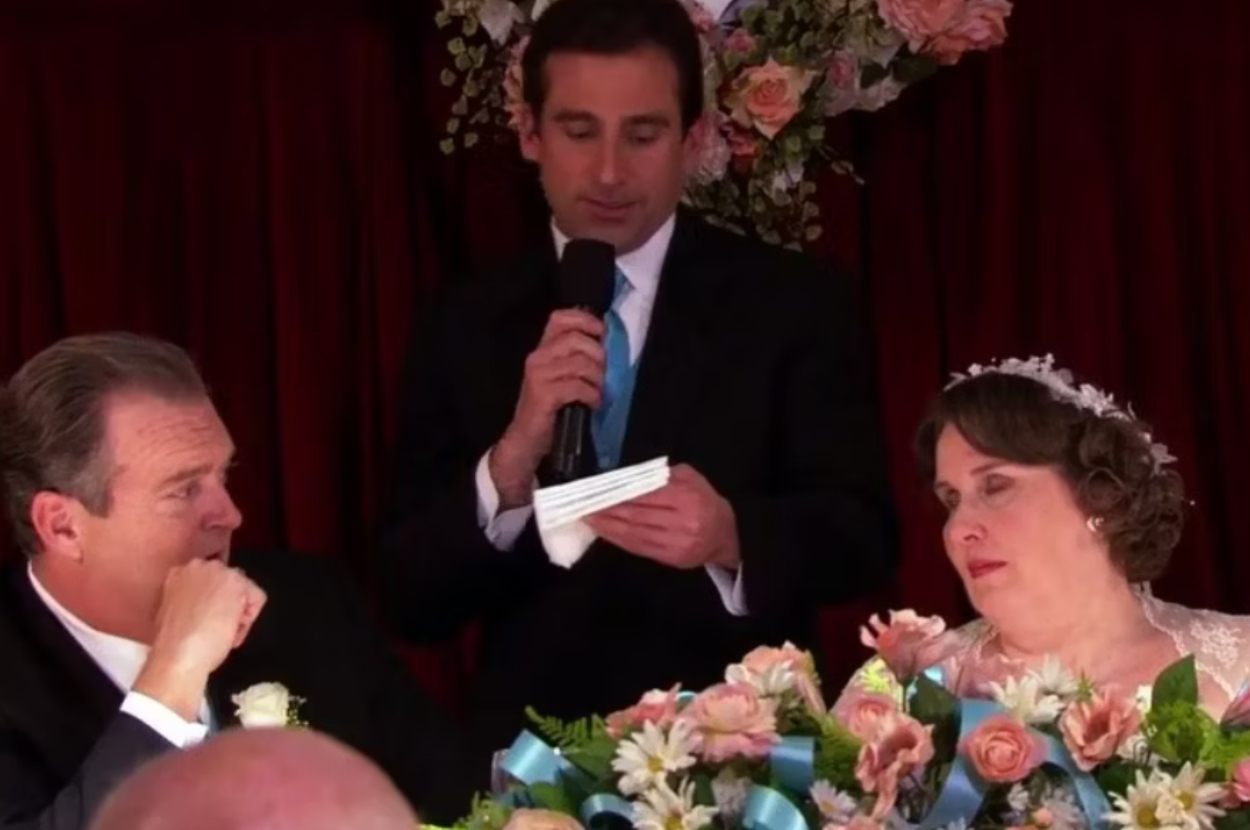 Steve Carell in a suit, holding a microphone, stands between Brian Baumgartner and Phyllis Smith in bridal attire, both seated at a decorated table