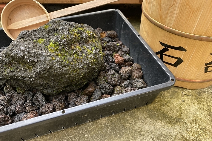 木製の桶と柄杓が置かれている場所に、黒い石や岩の断片が容器に入れられている写真。