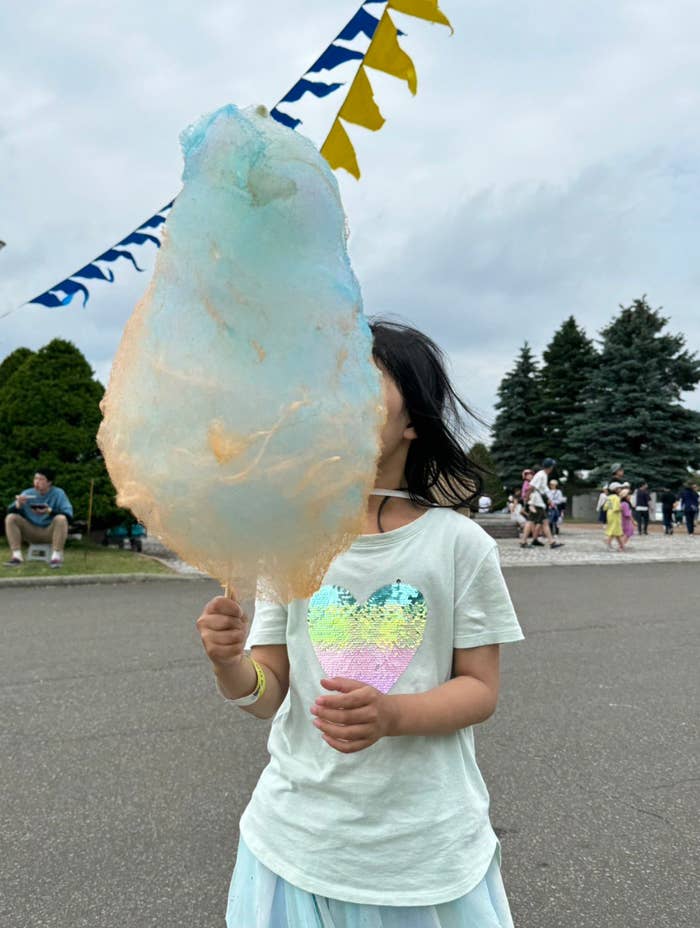 少女が心の形のシャツを着て綿菓子を持っています。背景には祭りのような場所で人々がいます。