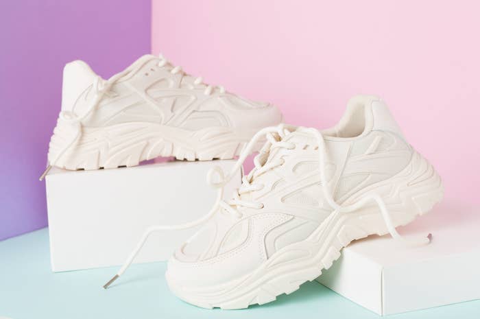 White chunky sneakers displayed with one shoe on top of a white box and the other next to it. The background has purple and pink sections