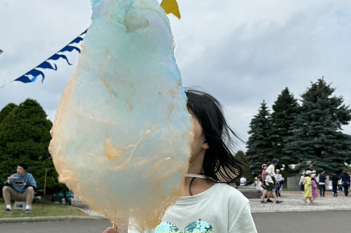 少女が心の形のシャツを着て綿菓子を持っています。背景には祭りのような場所で人々がいます。