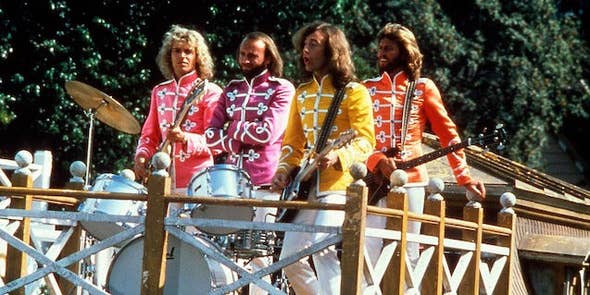 Four men, Barry Gibb, Robin Gibb, Maurice Gibb, and Colin Petersen, are playing music outdoors in colorful, decorative military-inspired jackets