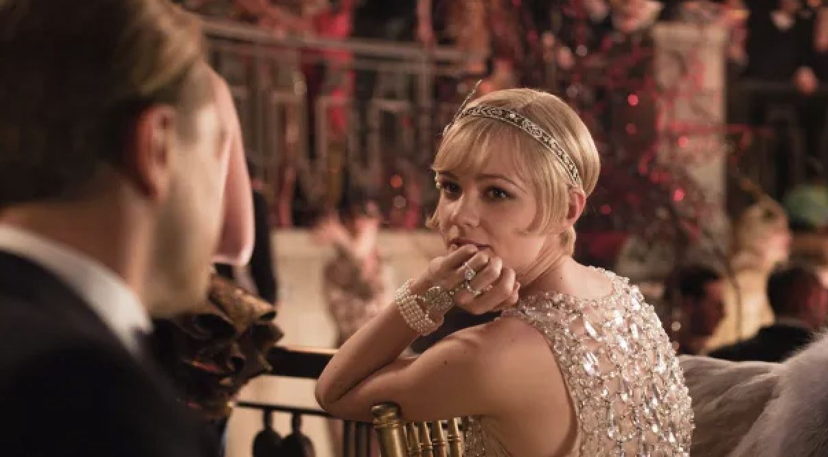 Carey Mulligan, in an embellished dress and headband, sits at a lavish party, looking over her shoulder at a man with his back to the camera
