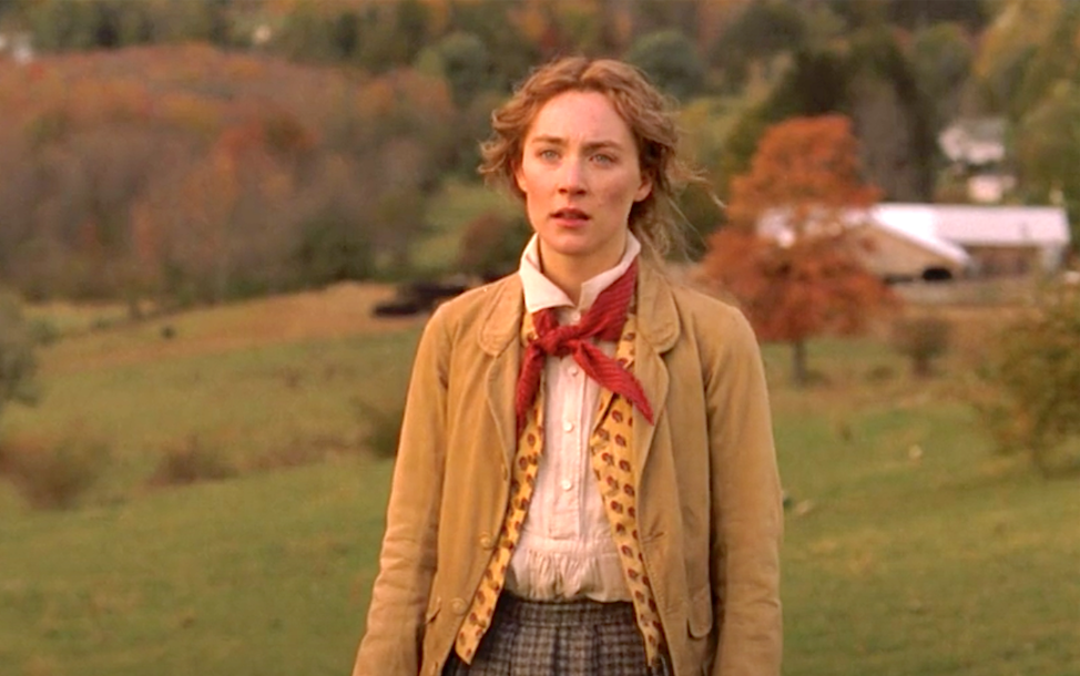 Saoirse Ronan in a pastoral setting, wearing a beige jacket, a red scarf, and patterned vest, standing on green grass with a background of trees and countryside