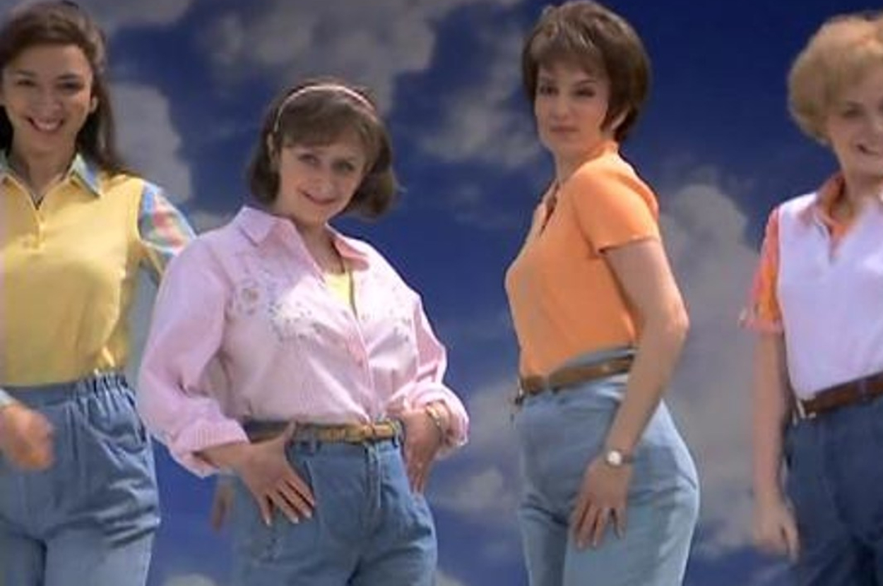 Four women, each smiling and posing in high-waisted jeans with hands on hips, stand against a sky backdrop. Clothing styles are from the 1980s