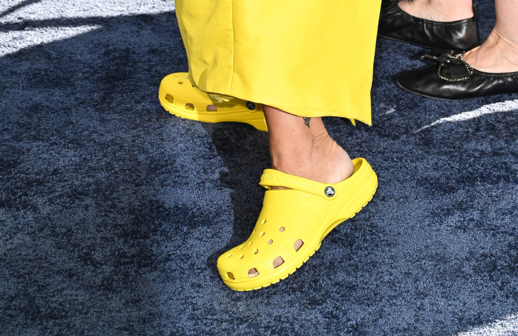 Two people standing; one in yellow Crocs with a small leg tattoo, and the other in black shoes with a jeweled buckle