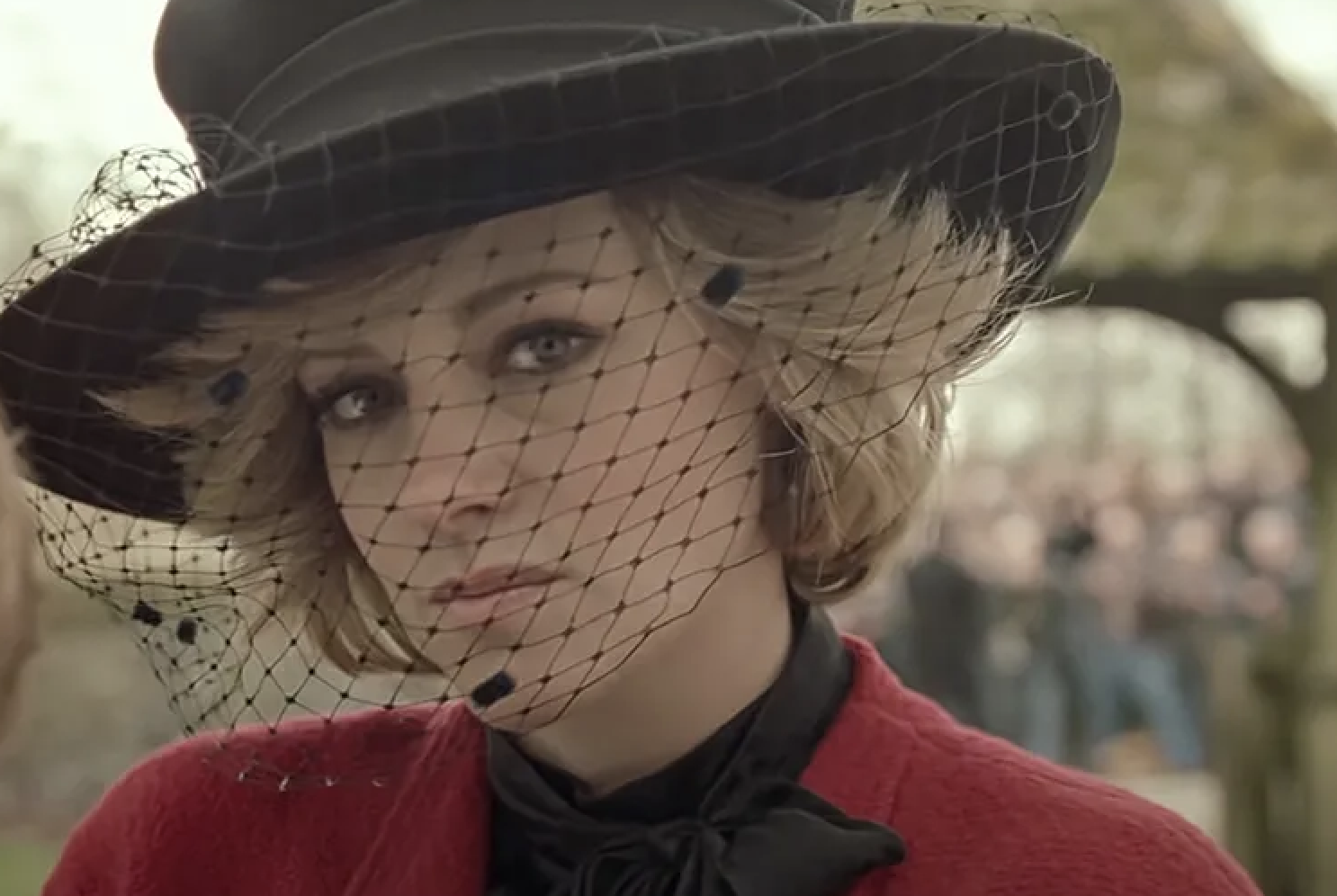 A woman dressed in 1980s-style clothing, including a black hat with a veil, looks intently at the camera