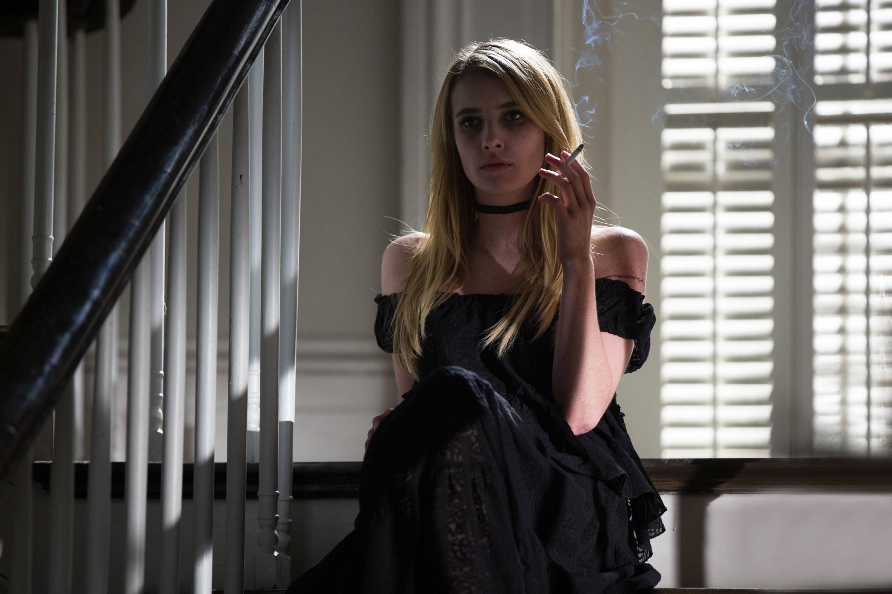 Emma Roberts sits on the stairs in an off-the-shoulder dress, holding a cigarette. She has long, straight hair and wears a choker necklace